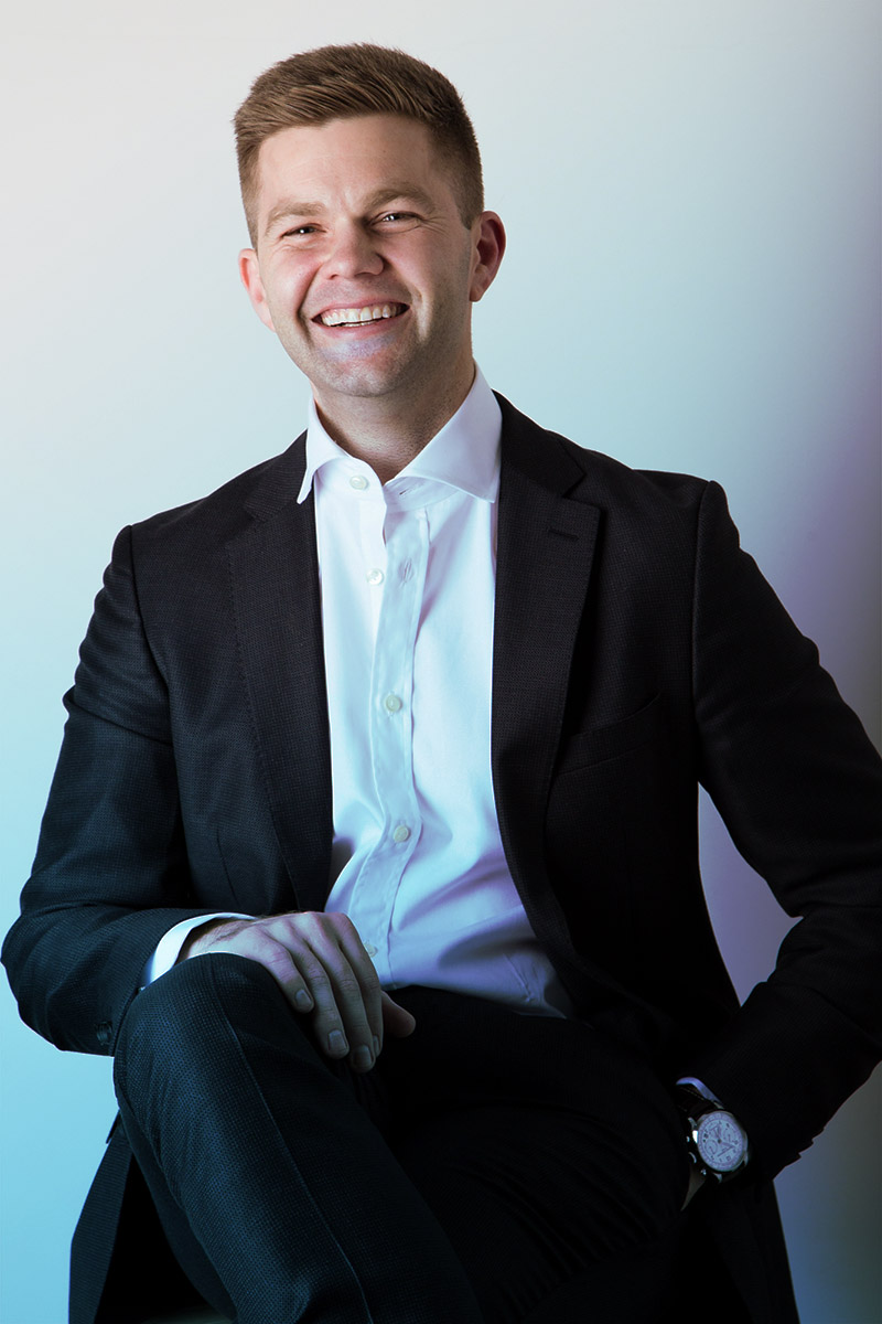 A smiling man in a black suit and white shirt, sitting and looking confidently at the camera.