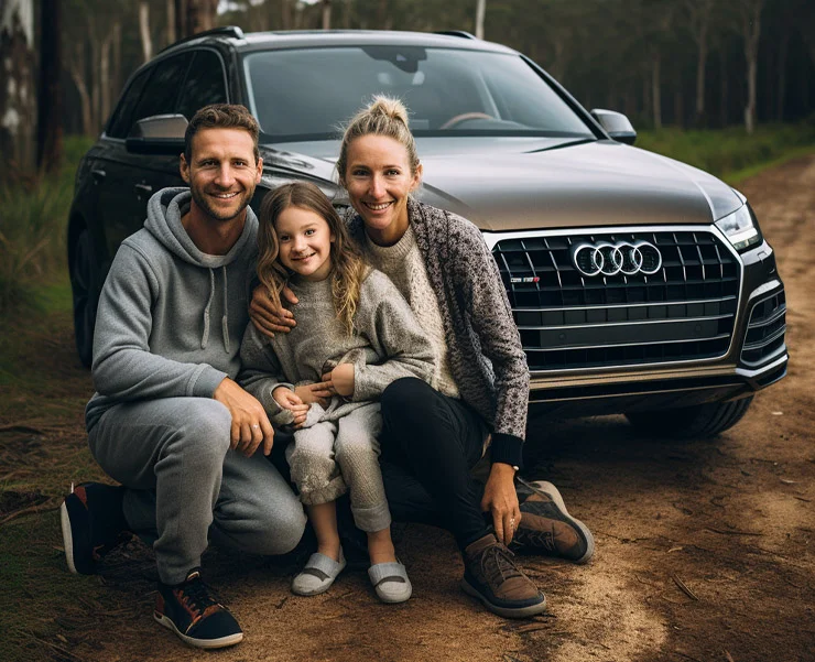 A smiling family of three, sitting together in front of an Audi SUV on a dirt road in a forest setting.