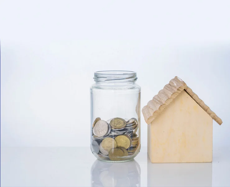 A jar filled with coins placed next to a small wooden house model on a white background.