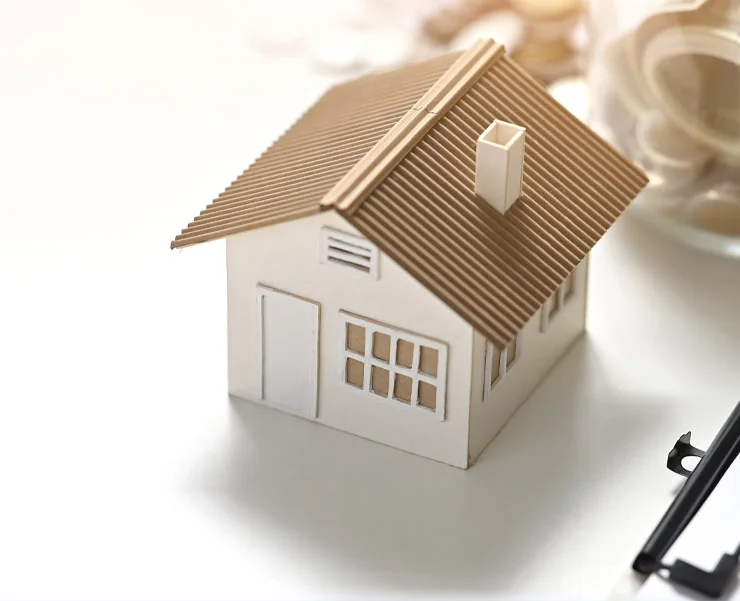 A small white model house with a brown roof placed on a white surface, with coins and a key visible in the background.