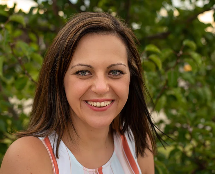 Smiling woman with shoulder-length brown hair, wearing a sleeveless top, standing outdoors with greenery in the background.