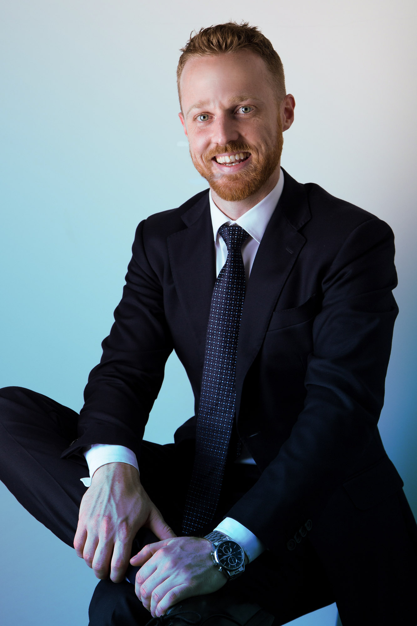 A man with short red hair and a beard, dressed in a black suit and tie, smiling confidently.