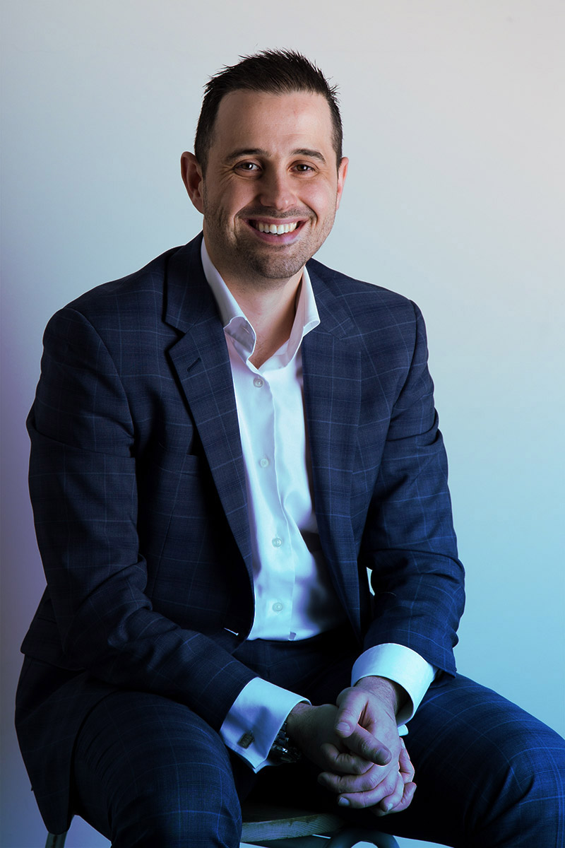 A man with short dark hair, dressed in a blue checkered suit and white shirt, smiling confidently.