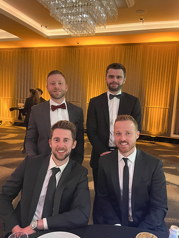 The Acton Wealth team dressed in black suits at the IFA Awards presentation, smiling for a group photo in a luxuriously decorated venue with a chandelier and golden curtains in the background.