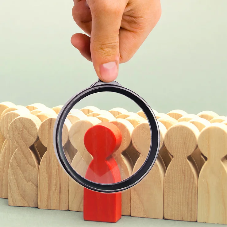 A magnifying glass held by a hand focuses on a red wooden figure among a group of natural wooden figures.