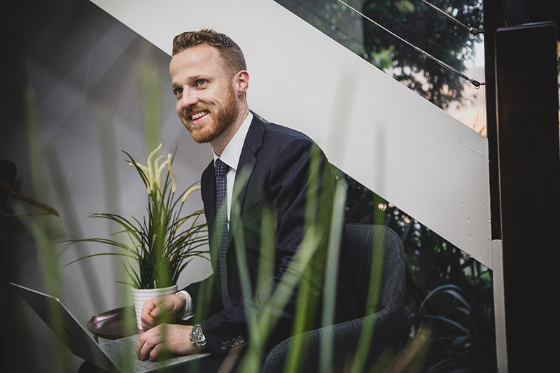 Blyth from the ActOn Wealth team, smiling and working on his laptop in an office setting, surrounded by indoor plants, exemplifying a professional yet relaxed working environment.