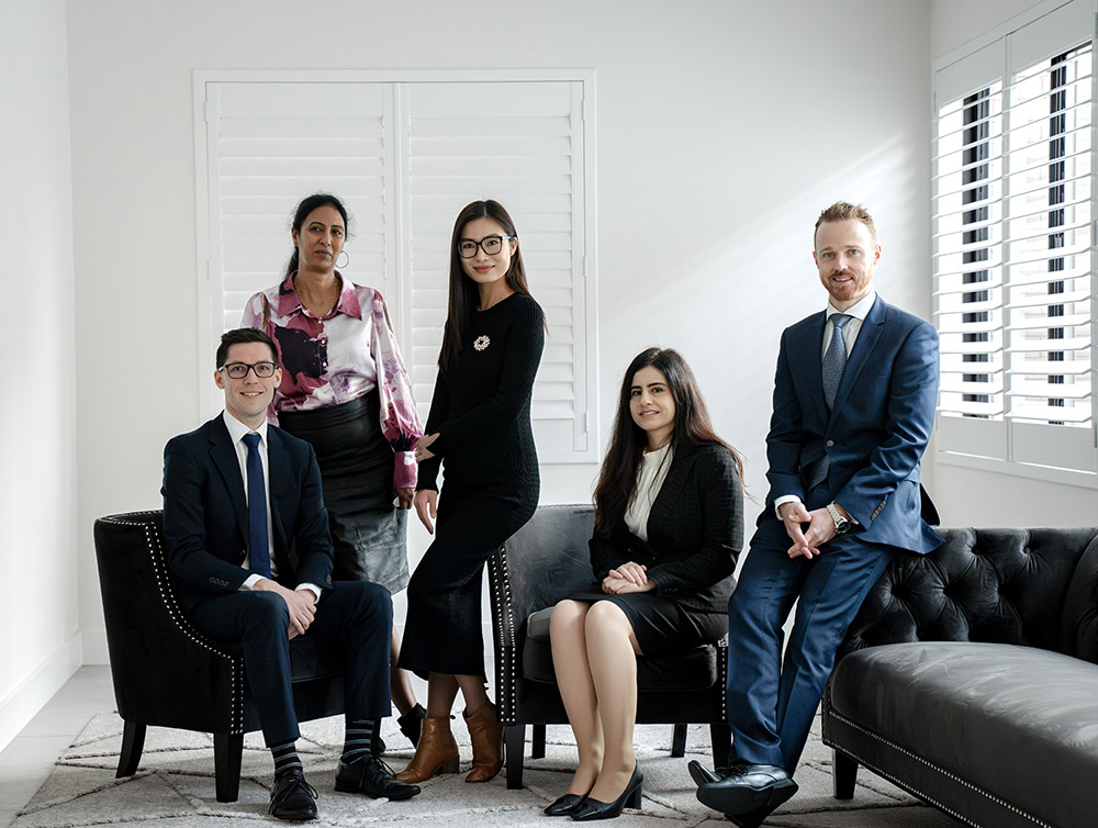 Members of the Actonwealth Lending Team posed in an office setting. From left to right: a seated young man in a suit, a standing woman in a floral blouse, a young woman in a black turtleneck and skirt, a seated woman in a grey suit, and a standing man in a blue suit, all smiling towards the camera.