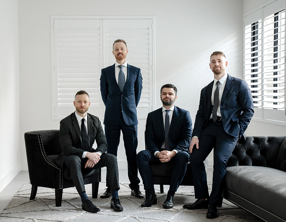 The Actonwealth Property Team, consisting of four well-dressed male professionals, confidently posing in a modern office setting with neutral decor and a large leather couch.