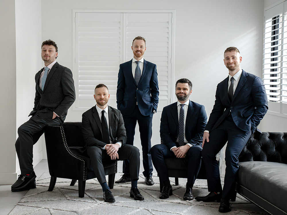 The ActOn Wealth Advice Team in their office, featuring five male financial advisors in suits. They are positioned with confidence around a modern setting with leather furniture and bright windows, projecting professionalism and approachability.