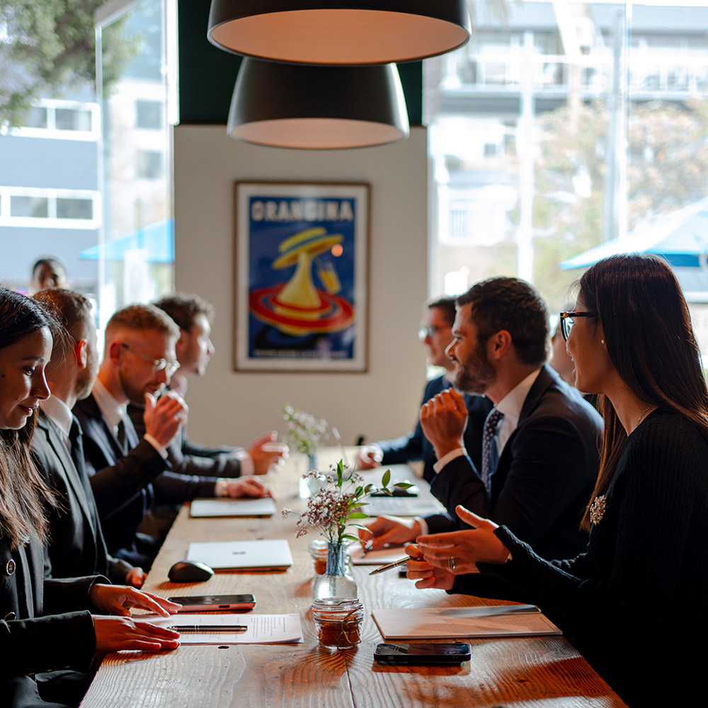 Members of the ActOn Wealth team engaging in a focused discussion during a work lunch at a modern cafe. They are seated around a long wooden table with laptops and documents, illuminated by a stylish overhead lamp. A vintage 'Orangina' poster decorates the background, adding a pop of color and charm to the setting.