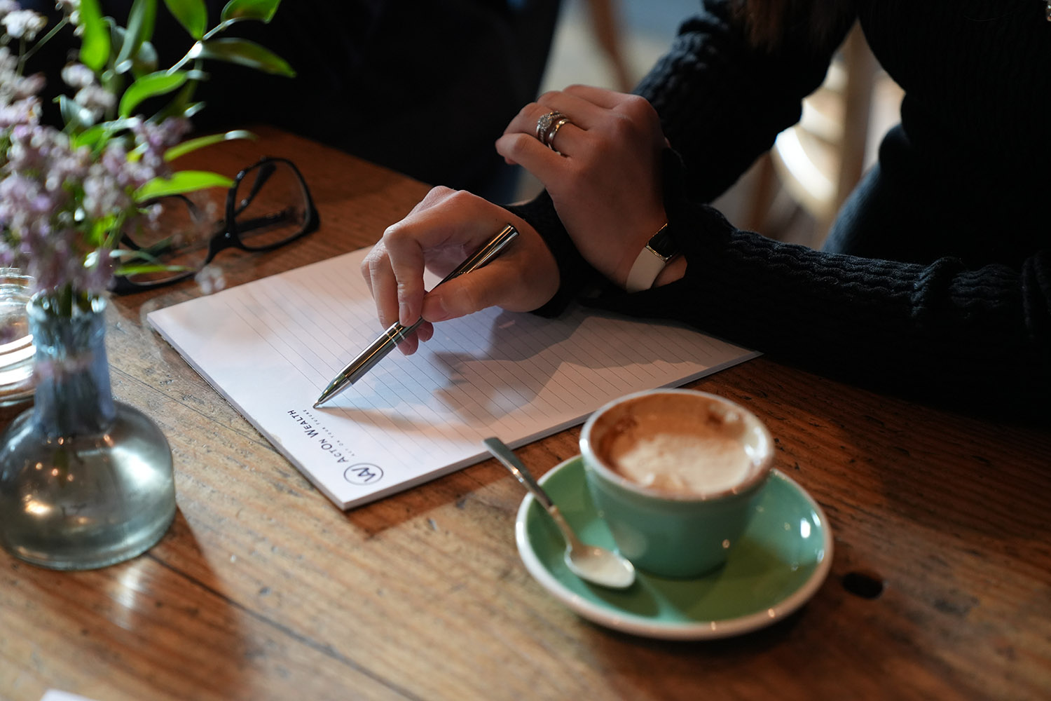 A person wearing a black sweater writing in a notebook at a table with a teal coffee cup and glasses next to a small vase of flowers, in a cozy café setting.