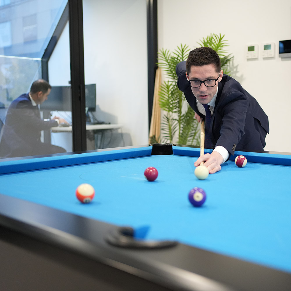 Man in a business suit playing pool, focusing on a shot, with another man working at a desk in the background in a modern office environment.