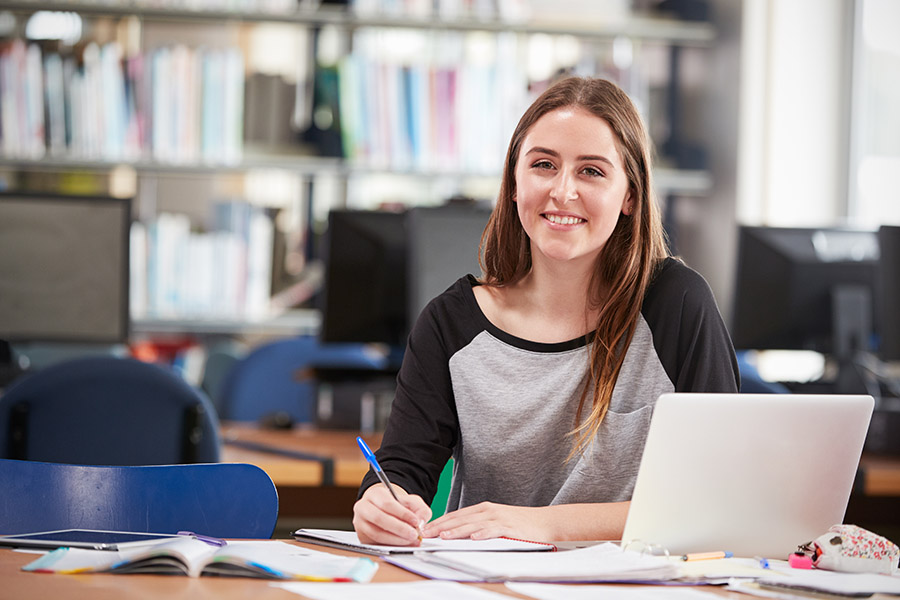 A young woman sitting in a library, smiling while working on her laptop and taking notes with a pen.