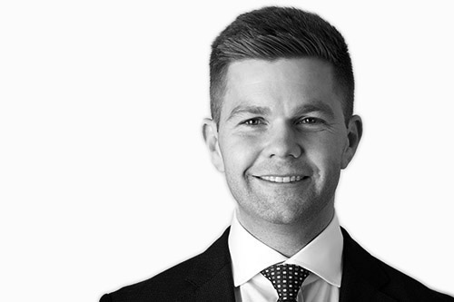 Portrait of Shannon Cramer, a young Caucasian male with short, neatly styled hair, wearing a formal dark suit and tie. He is one of the directors at Actonwealth, depicted in a black and white headshot with a confident smile.