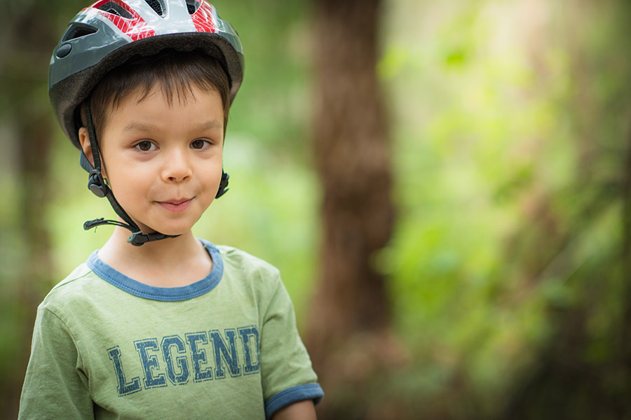 A young boy wearing a bicycle helmet and a green t-shirt with the word 'Legend' printed on it, standing outdoors with a blurred green background.