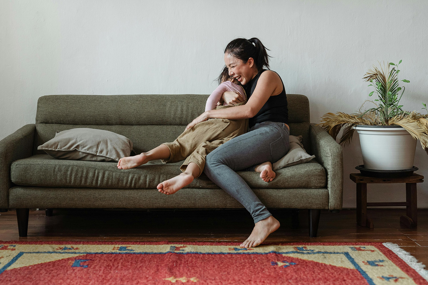 A joyful moment as a woman sits on a couch, playfully embracing her child in a warmly decorated living room. They are both smiling and engaging in a close, affectionate moment. The room features a green sofa, a large potted plant, and a vibrant rug, creating a cozy and inviting space.