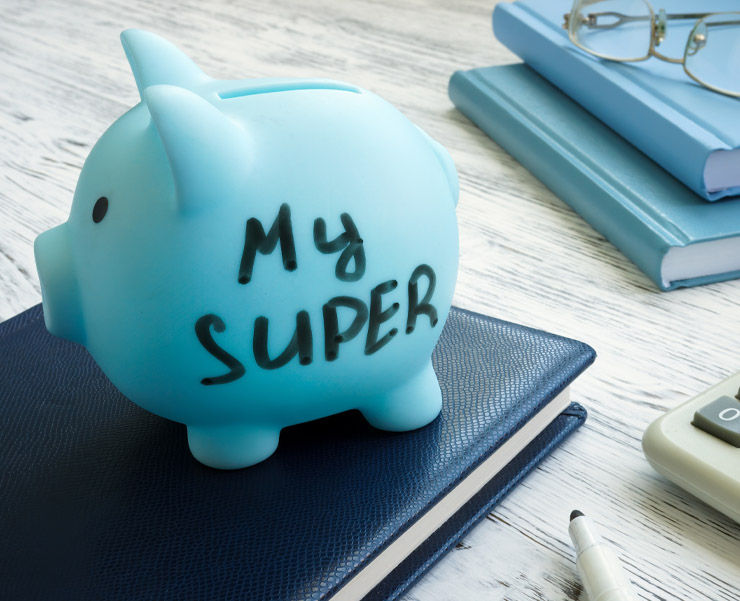 A blue piggy bank with 'My Super' written on it, resting on a navy blue diary beside a calculator and glasses on a wooden table.