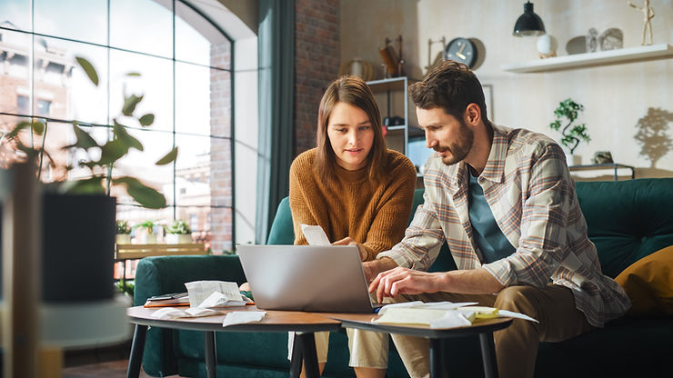 A couple sitting on a couch working on a laptop, surrounded by papers and receipts in a cosy living room.