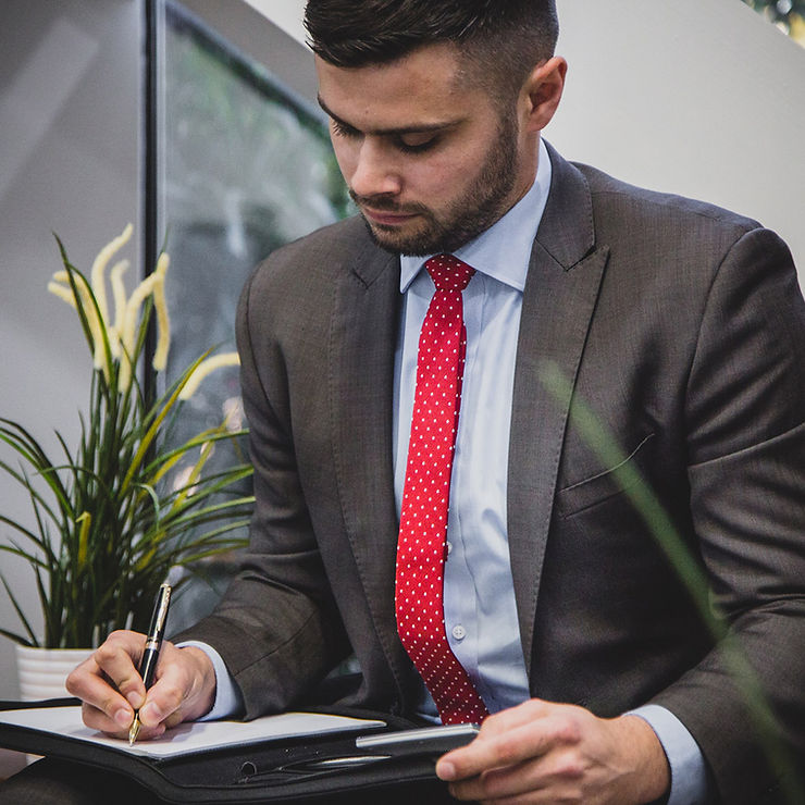 A man in a suit and red tie writing in a notebook while seated in a professional setting.