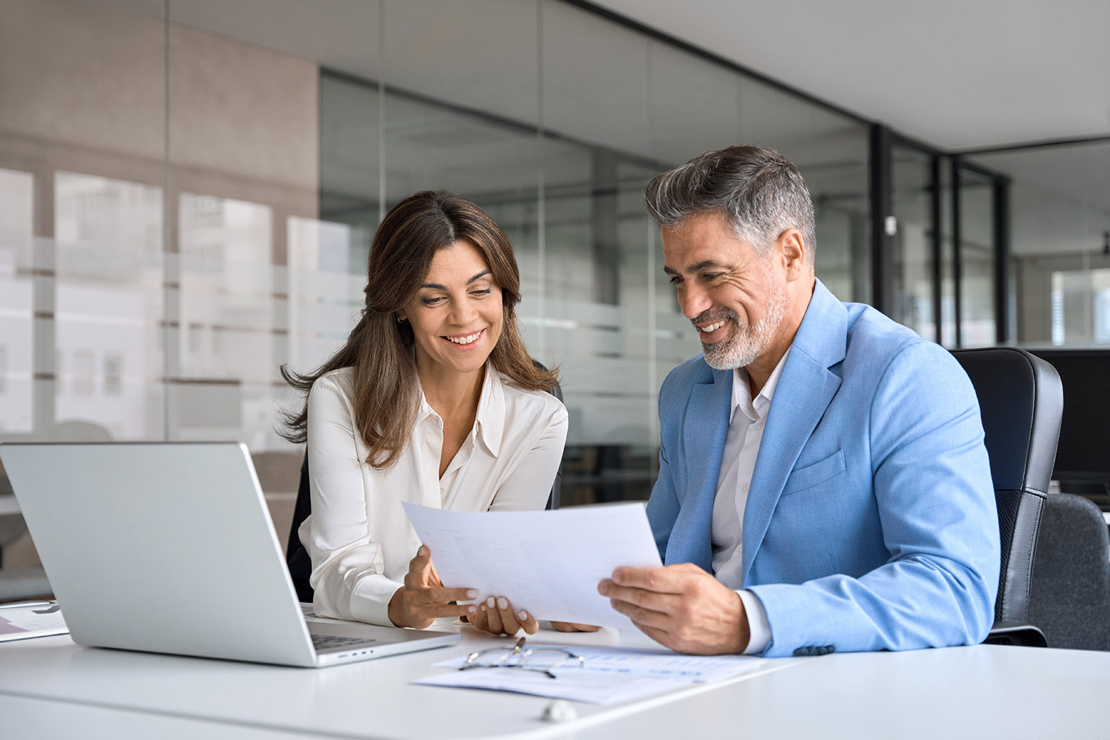 Two professionals, a man and a woman, smiling while reviewing a document together in a modern office setting.