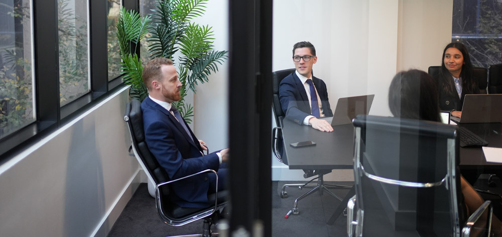 A professional meeting in progress at the ActOn Wealth office, featuring four male team members in business attire, engaged in discussion around a conference table with laptops and documents.