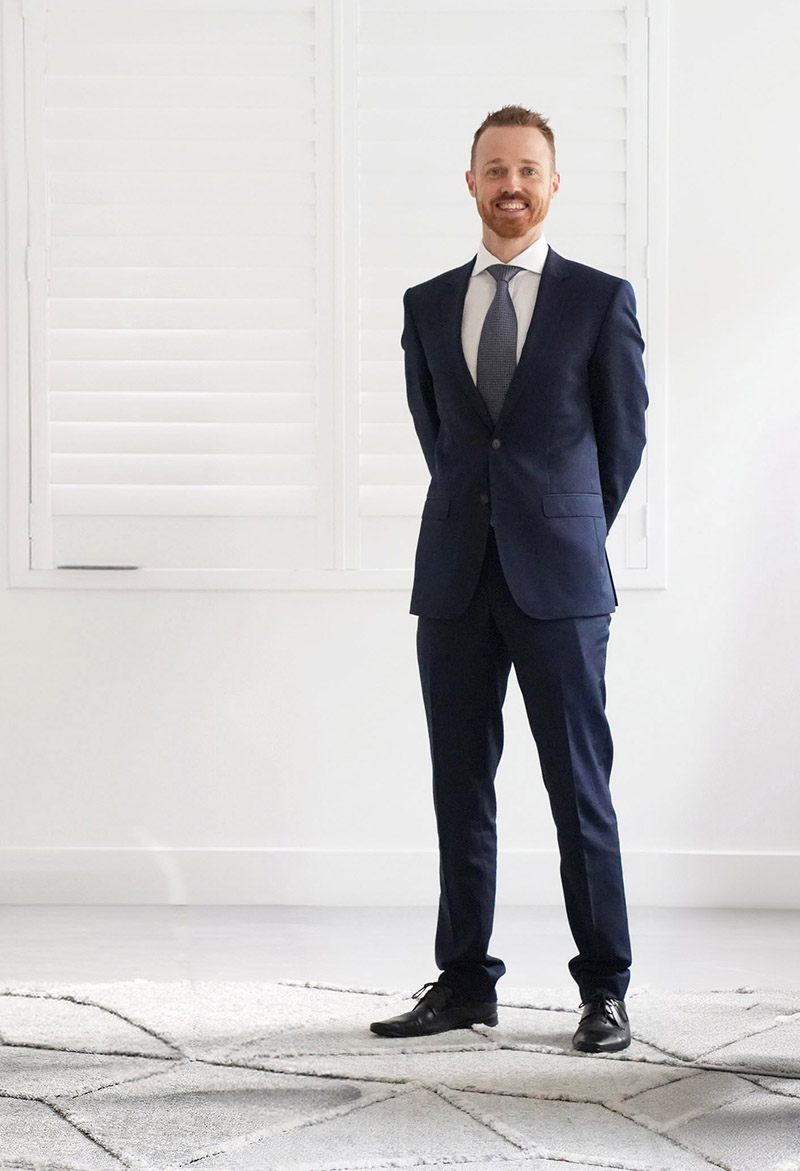 A man in a navy blue suit standing in a bright, modern office with his hands behind his back, smiling at the camera.