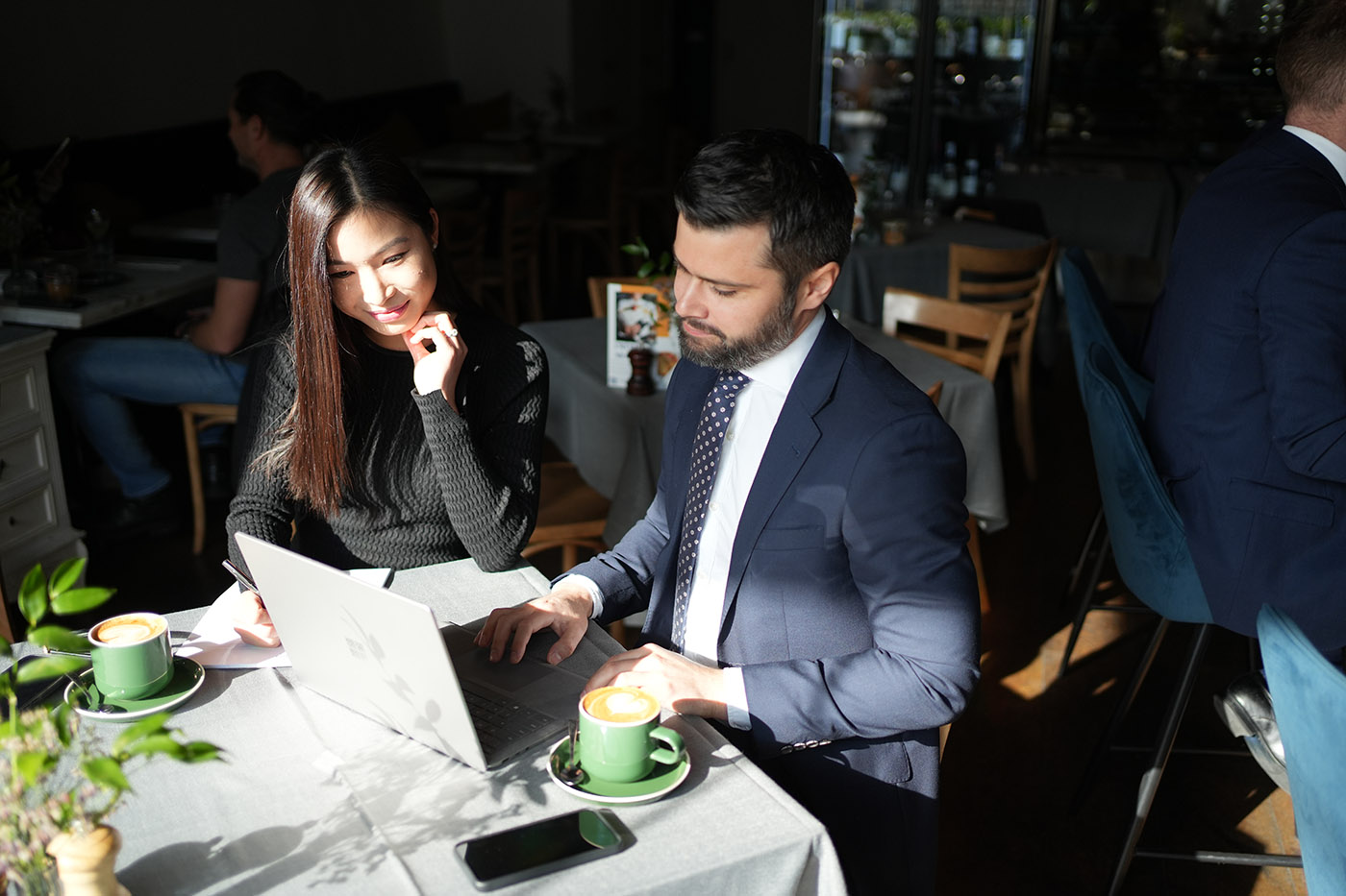 A man in a suit and a woman in a black sweater working together on a laptop in a café, with coffee cups on the table.